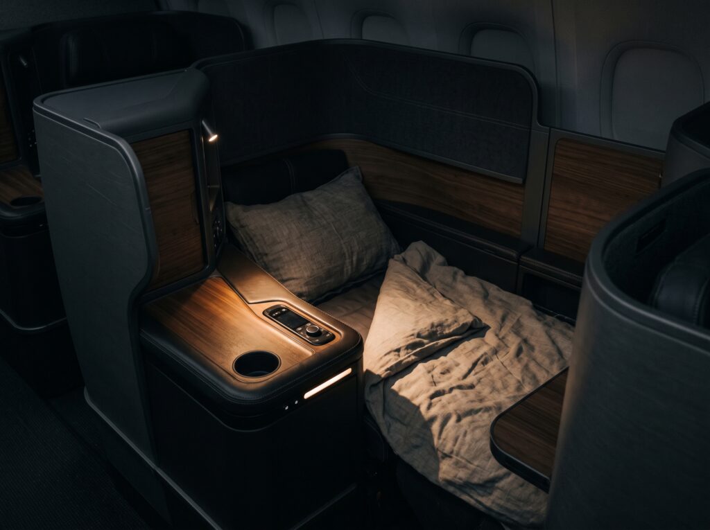 Close-up of an enclosed business-class seat in shadow with a warm highlight on the armrest and a lie-flat bed dressed in linen and wood panels.